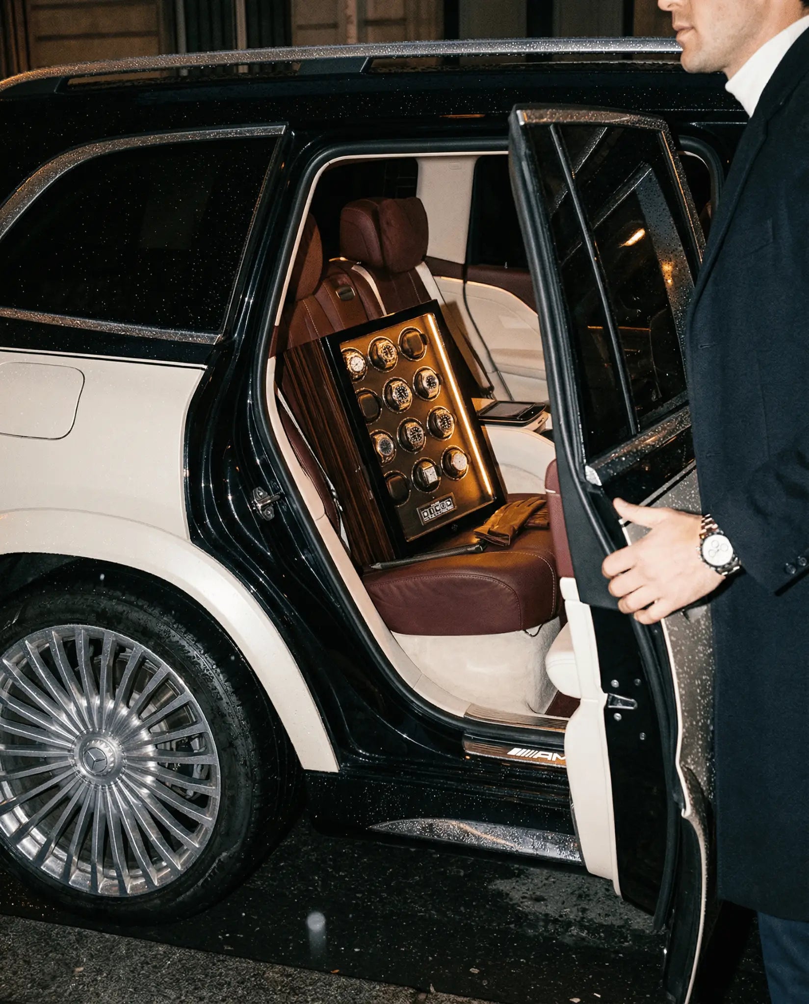 Remontoir montre automatique Mercier & Fils Mazarin XII pour 12 montres garni de montres de luxe, posé sur la banquette cuir bordeaux d'un Mercedes GLS Maybach, homme en costume ouvrant la portière de nuit sous la pluie